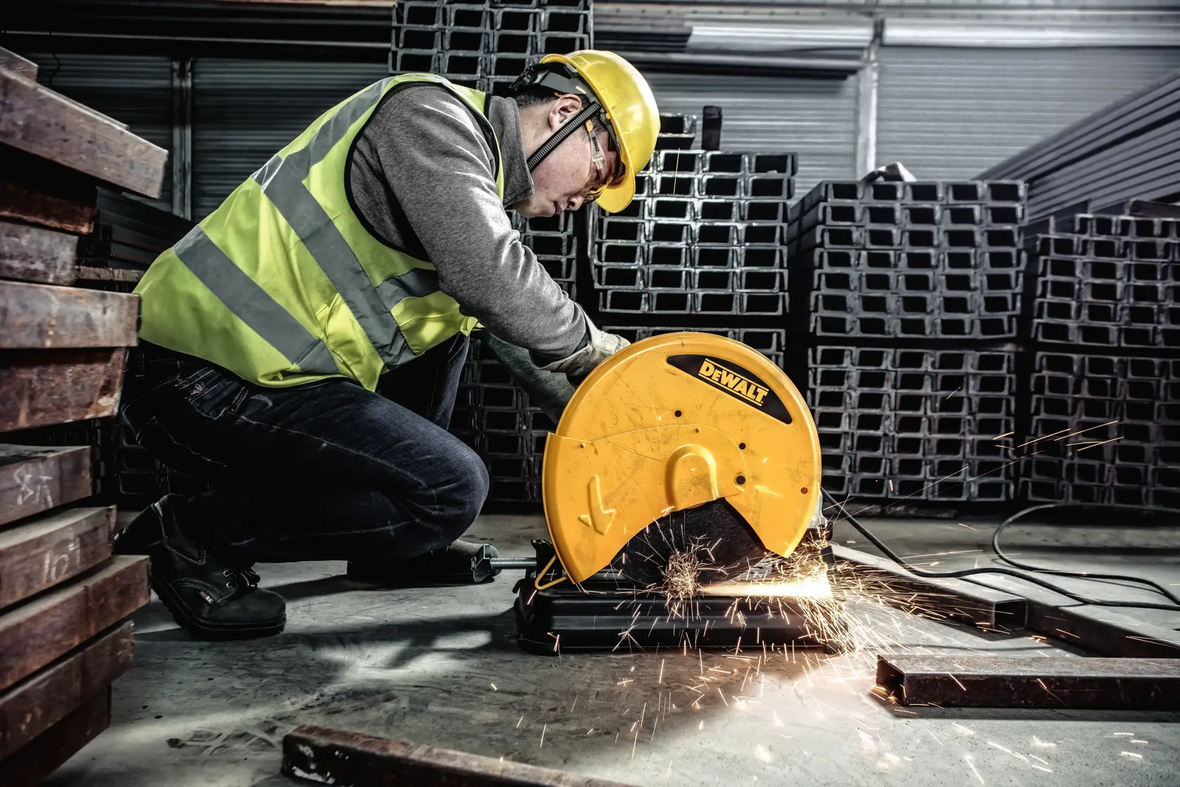 A person wearing a yellow safety helmet and reflective vest uses a DEWALT metal cutting saw to cut steel bars in an industrial warehouse. Sparks are visible during the cutting process.