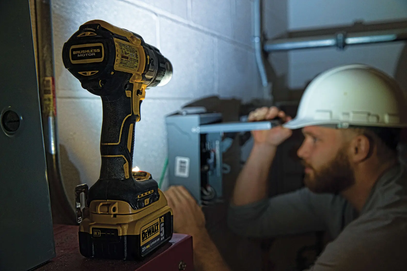 A person wearing a green safety jacket and white hard hat is using a DEWALT DCD996P2 cordless power drill to work on a concrete wall in an industrial setting. The face is obscured for privacy.