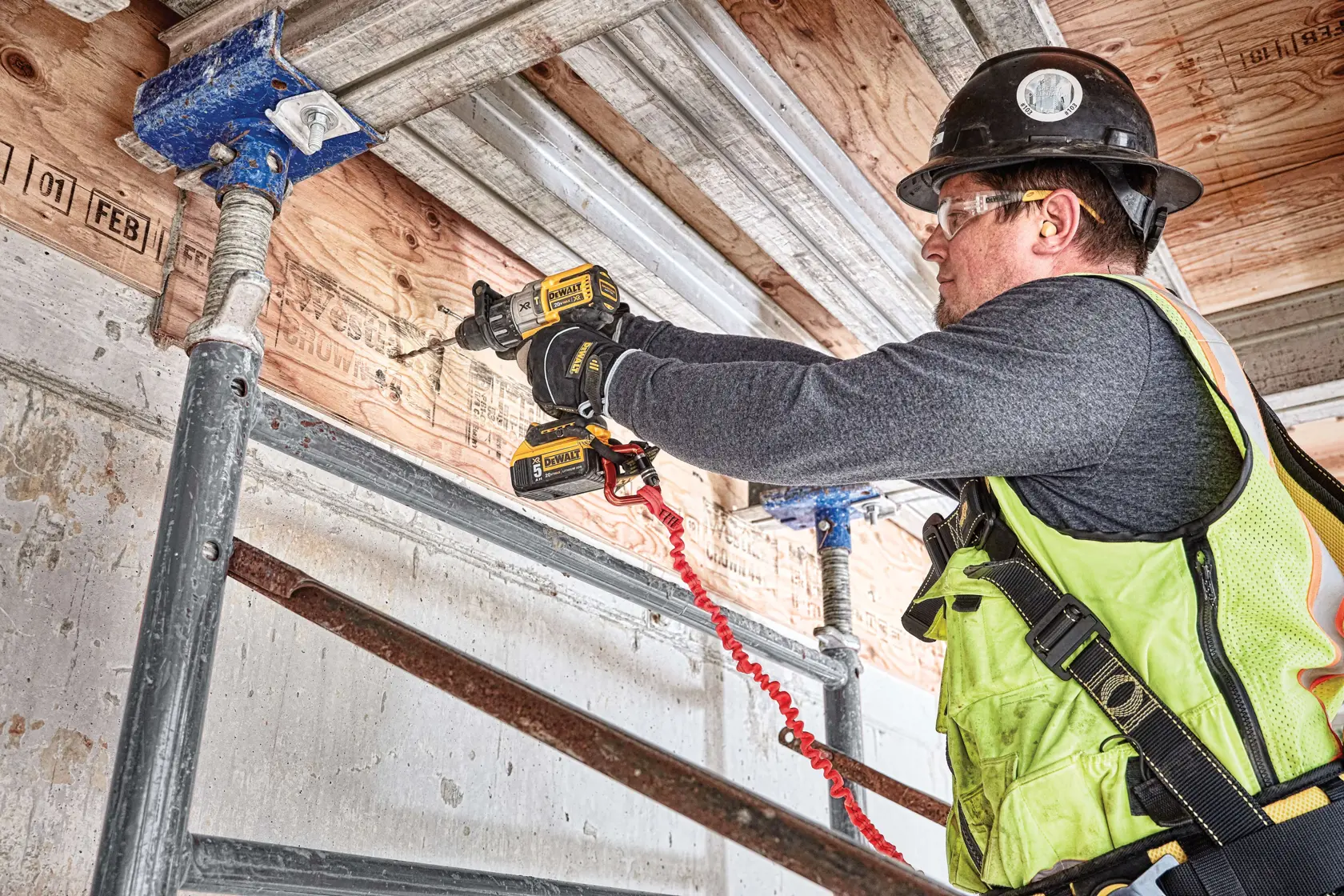 A person using a DEWALT DCD996P2 power drill to drill through a wooden beam at a construction site. There is sawdust on the floor and the person is wearing work boots and jeans.