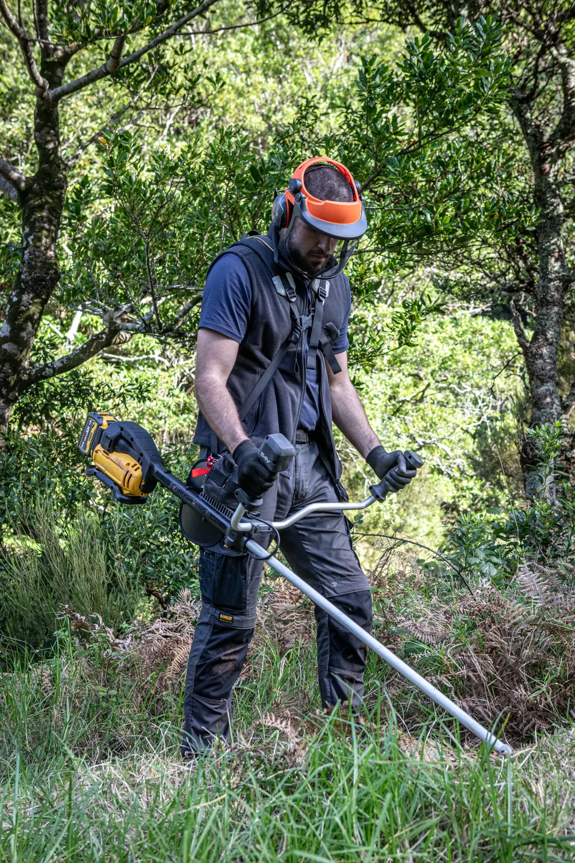Person uses DEWALT brush cutter DCMST922N, DCMST922N-XJ in grassy area with safety gear outdoors.