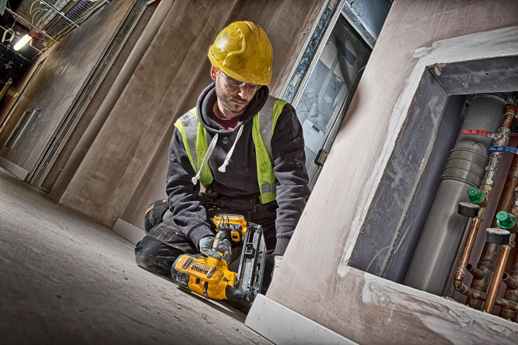 Worker kneeling indoors using a DEWALT DCN660D2 nailer on baseboards. Wearing a yellow hard hat and safety vest.