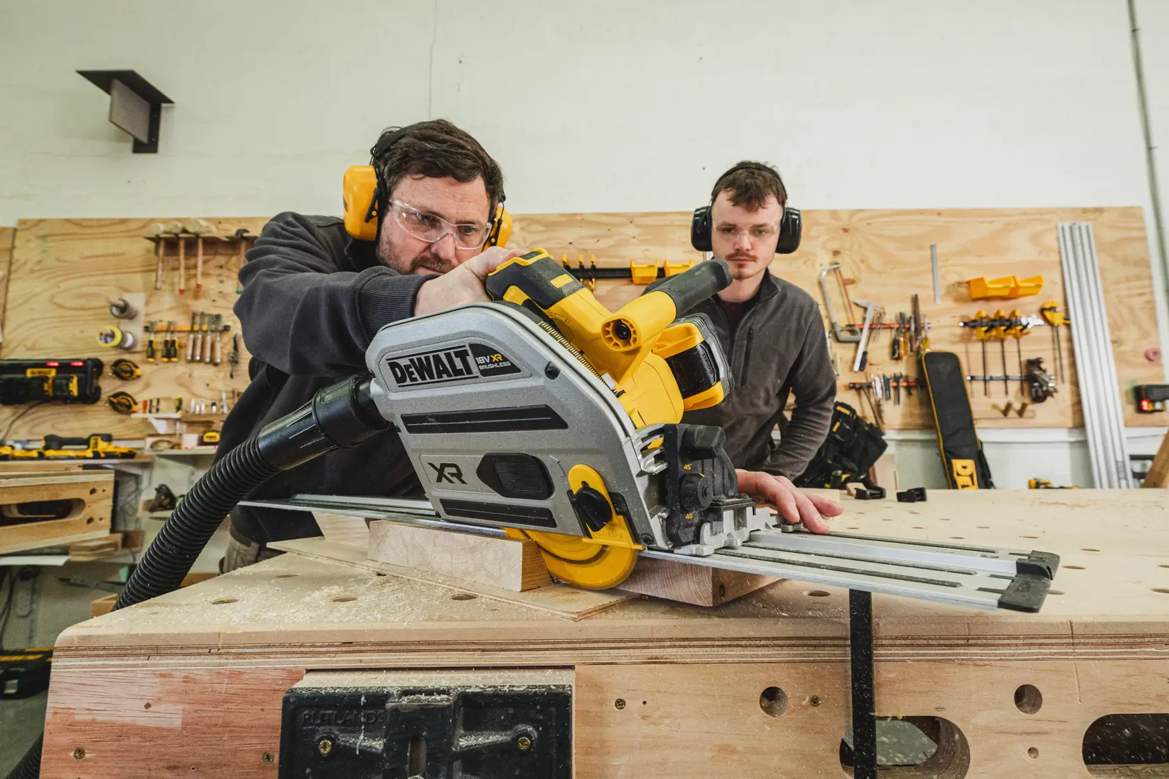 Two people using a DEWALT cordless track saw to cut wood in a workshop with tools on the wall.