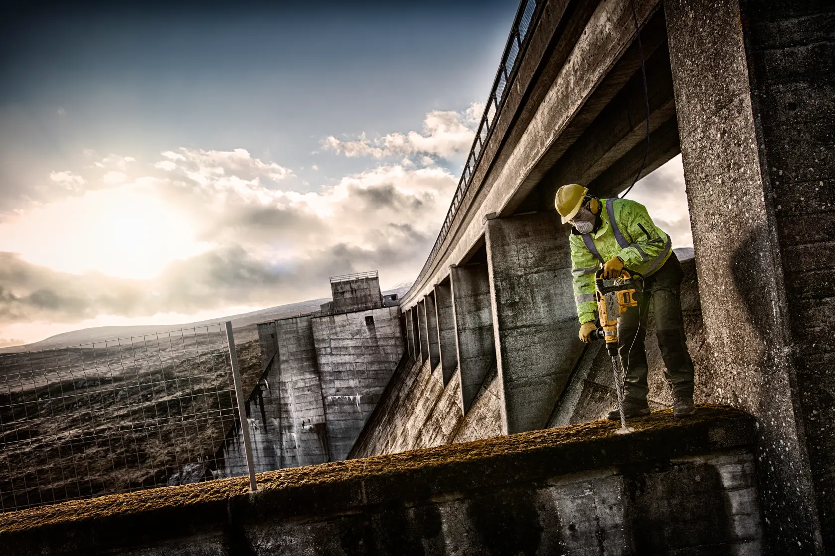 Arbeiter nutzt DEWALT-Bohrer auf Beton bei einem Damm im Freien bei Sonnenuntergang.
