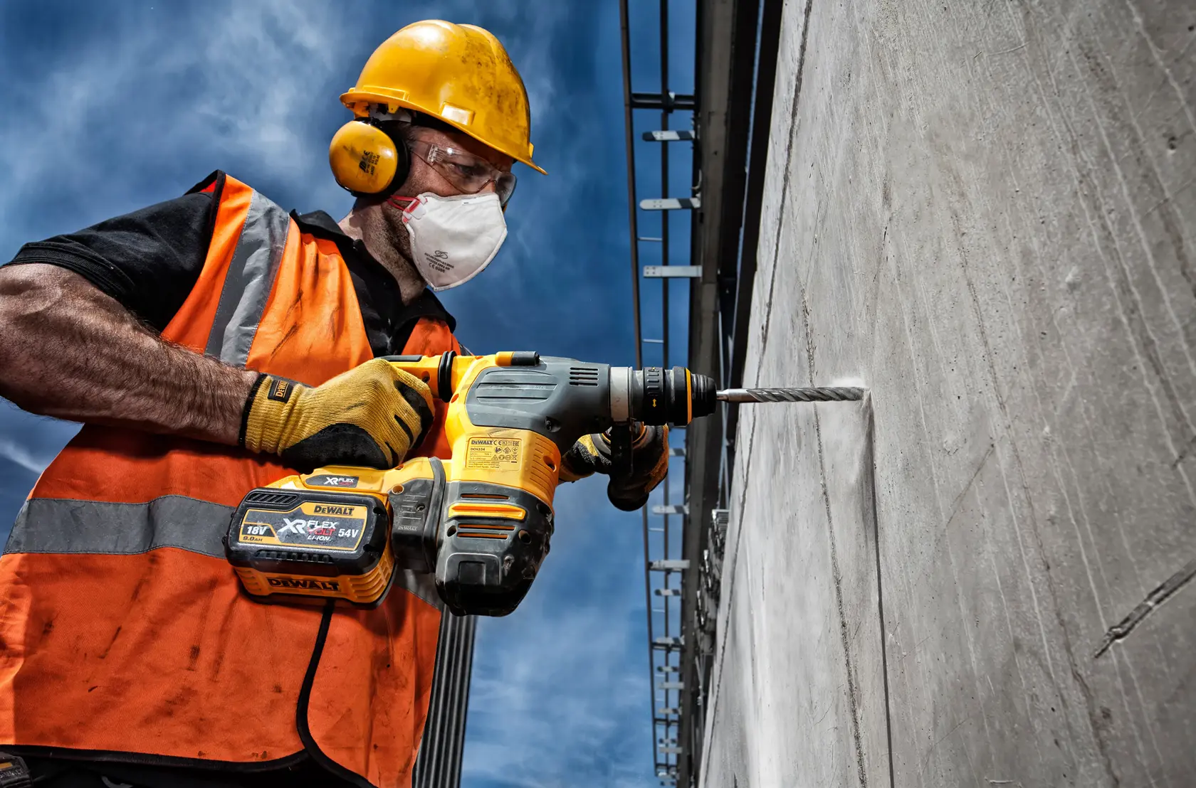 Trabajador perfora pared de concreto con taladro DEWALT al aire libre, usando equipo y chaleco naranja.