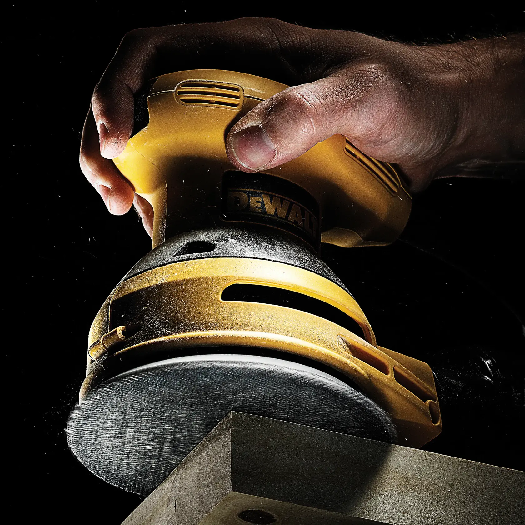 Hand mit DEWALT Schleifer beim Schleifen von Holz. Nahaufnahme, Staub sichtbar, schwarzer Hintergrund.