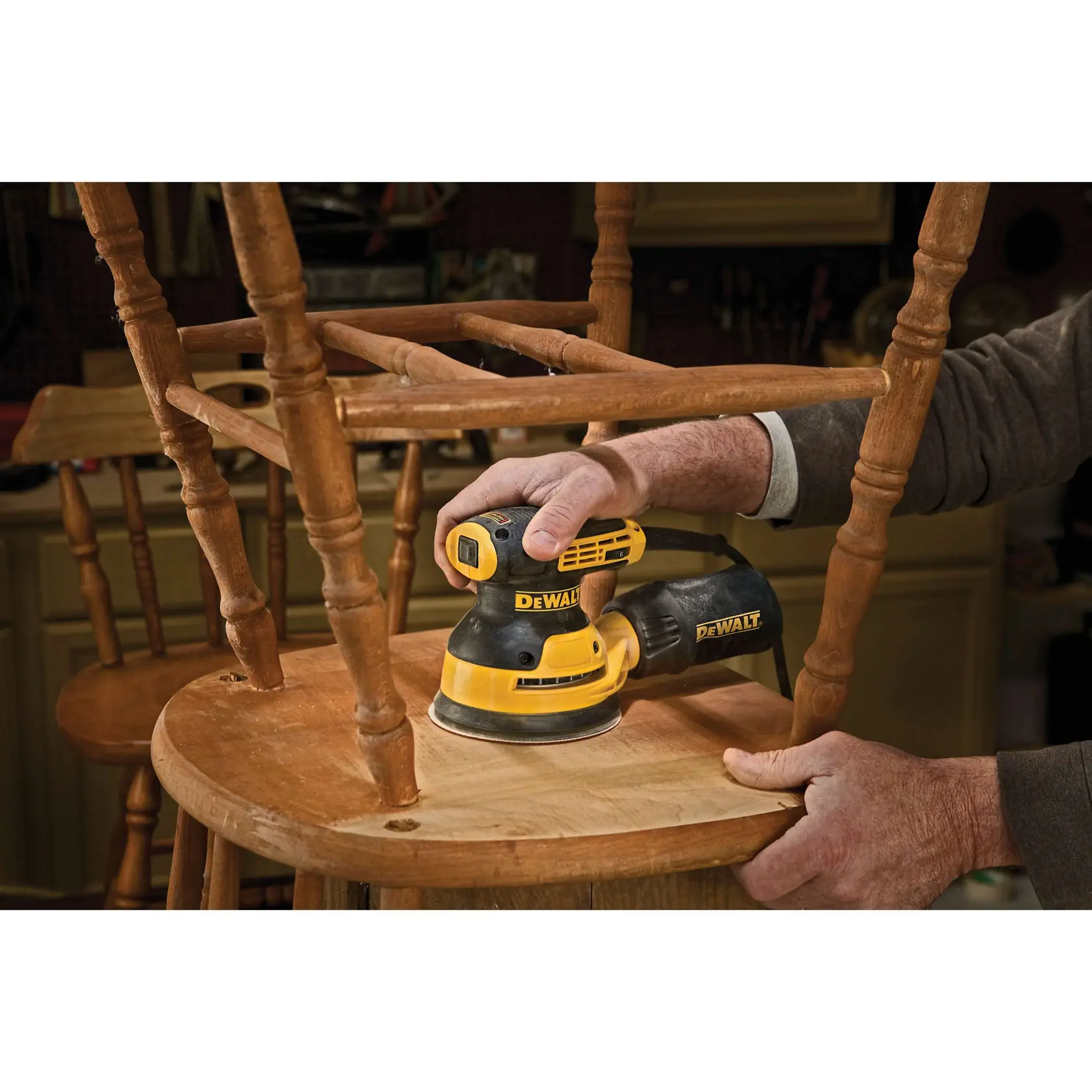 A person using a DEWALT DWE6423 random orbital sander to sand a wooden door in a woodworking shop. Some metal hardware pieces are positioned on the table nearby, and the person is wearing a protective mask.