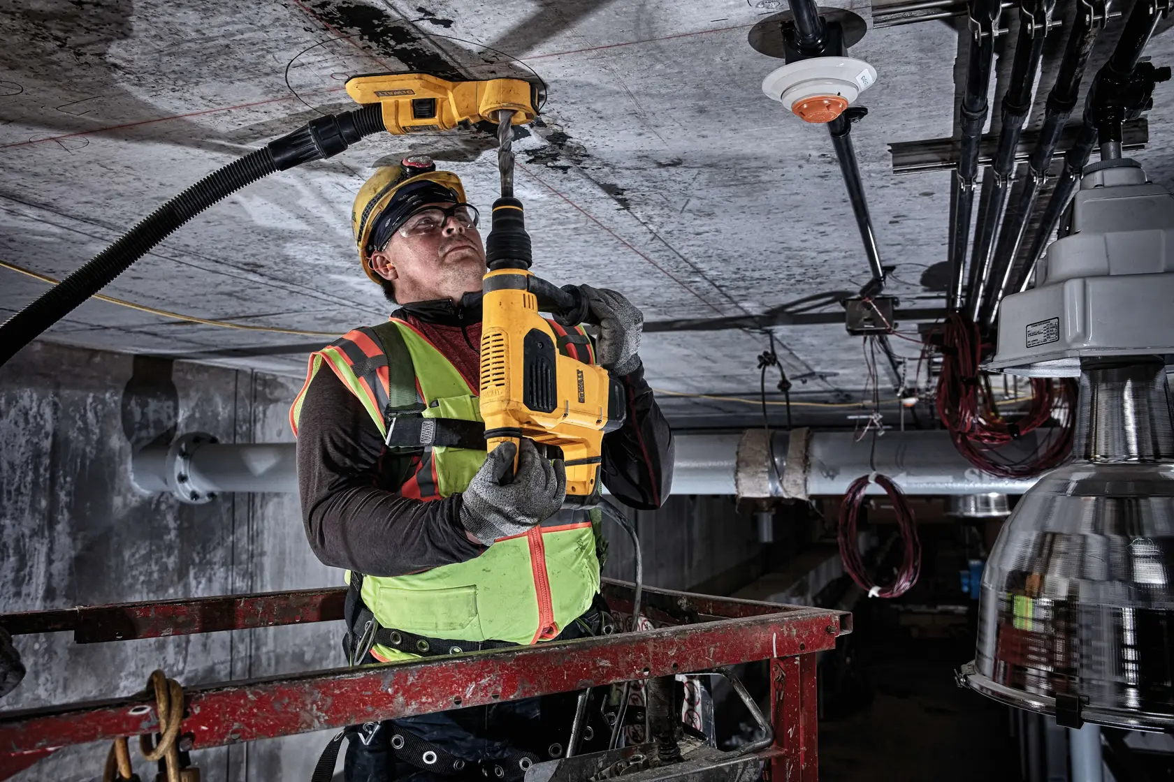 A worker wearing a green safety vest and helmet is using a DEWALT DWH050 dust extraction system attached to a yellow drill, drilling into a concrete ceiling in an industrial setting.