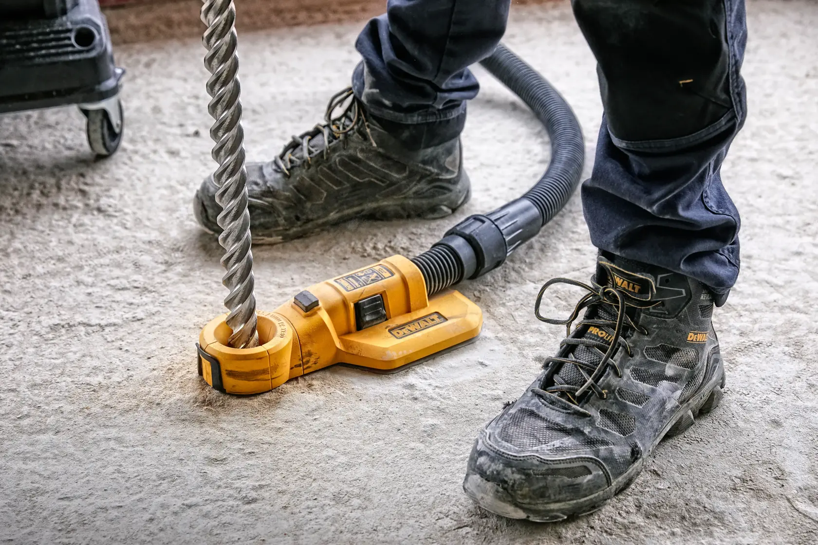 A close-up of a DEWALT DWH050 dust extraction attachment connected to a large drill bit and hose, being used on a concrete surface by someone wearing safety boots and work pants.