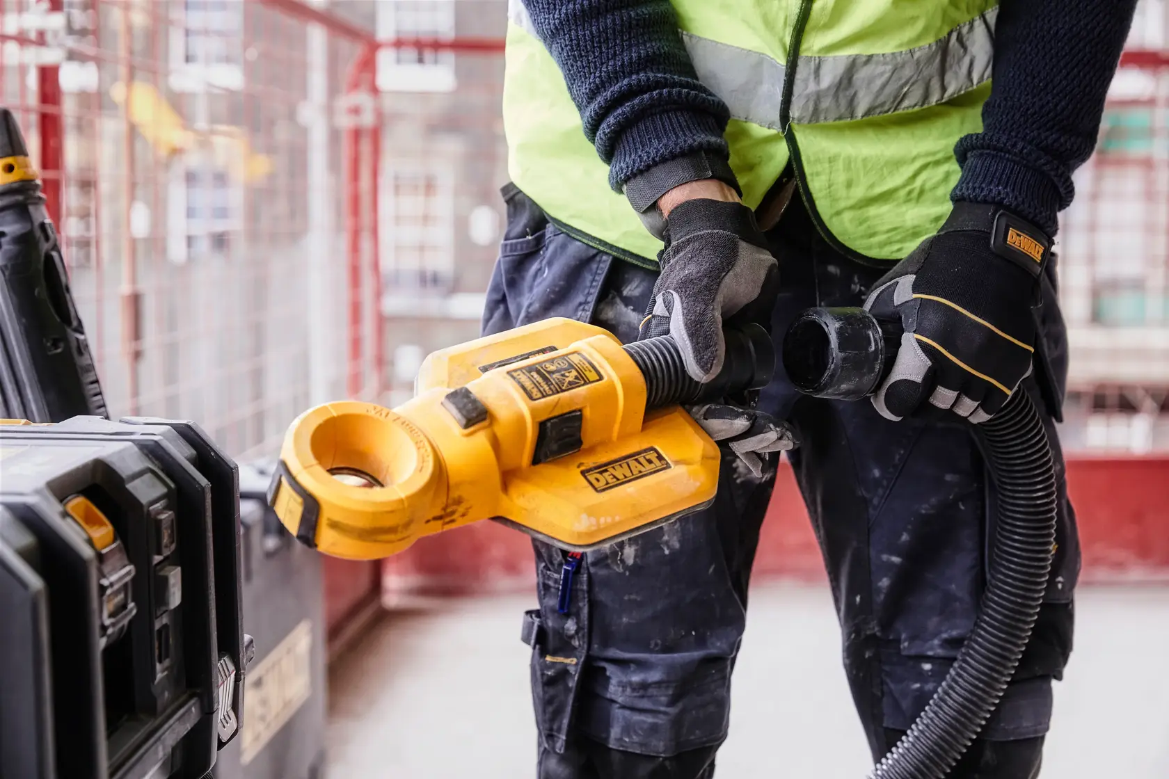 A person wearing safety gloves and a reflective vest uses a DEWALT DWH050 tool attachment for dust collection at a construction site.