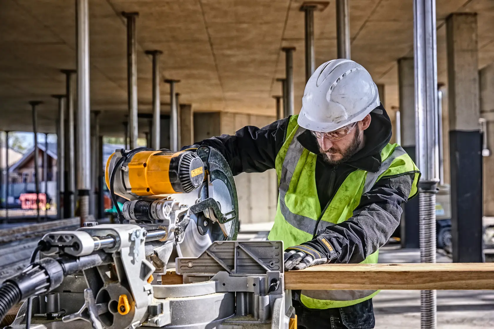 A DEWALT DWS780 miter saw is being operated by a worker wearing safety gear and a helmet at a construction site. The saw is cutting a piece of wood.
