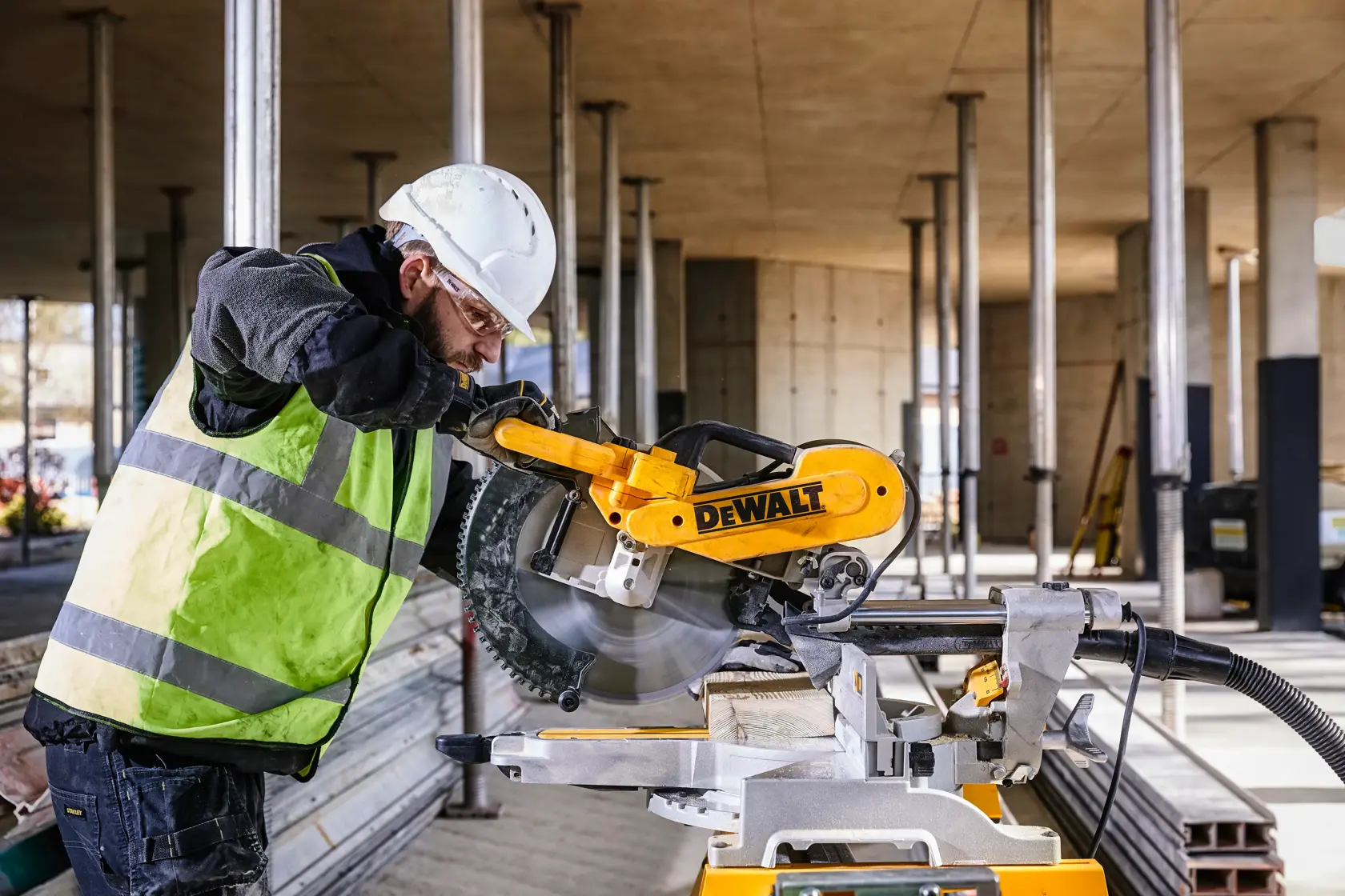 A person wearing a hard hat and safety vest is operating a DEWALT DWS780 miter saw at a construction site with metal supports and building materials visible in the background.