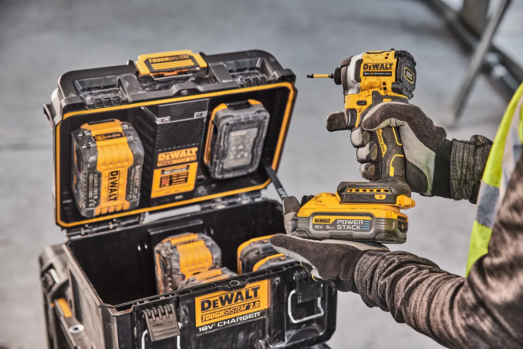 Person holding DEWALT drill, charging case with batteries and tools visible. Industrial setting.